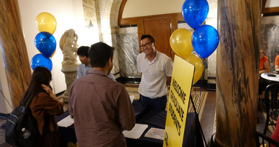 Graduate College representative welcome two new graduate students at orientation check-in table with balloons and welcome sign